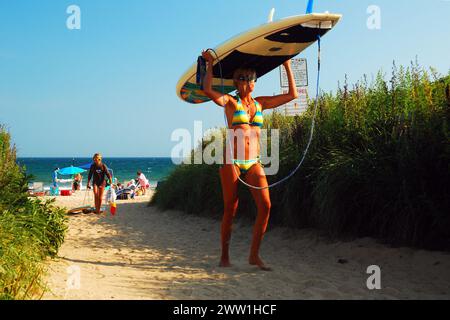 Eine starke junge Frau trägt ihr Surfbrett ausgewogen auf dem Kopf, während sie vom Strand zurückgeht Stockfoto