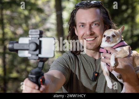 Ein Mann benutzt ein Handy-Gimbal, um sich selbst zu filmen und einen chihuahua, den er hält. Der Film spielt in einem natürlichen Waldgebiet. Stockfoto