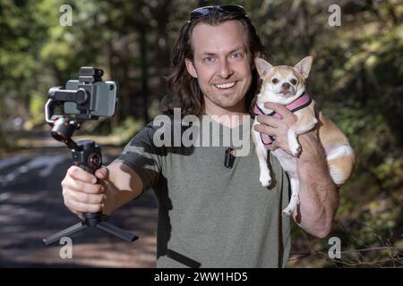 Ein Mann benutzt ein Handy-Gimbal, um sich selbst zu filmen und einen chihuahua, den er hält. Der Film spielt in einem natürlichen Waldgebiet. Stockfoto