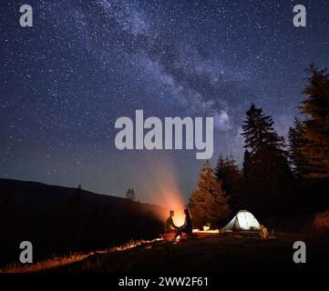 Nächtliches Camping in den Bergen unter himmlischem Baldachin. Mann und Frau Wanderer entspannen sich am flackernden Feuer und Zelt inmitten der sylvan-Gelassenheit, unter der ätherischen Schönheit des Chumack Trail. Stockfoto