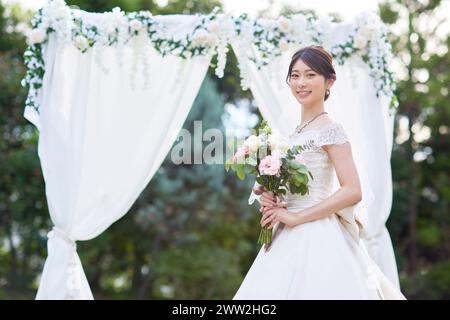 Asiatische Braut im Hochzeitskleid, die vor einem weißen Bogen steht Stockfoto