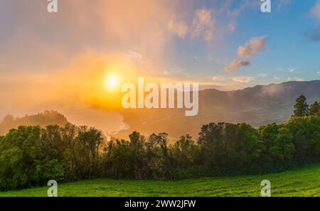 Genießen Sie den ruhigen Sonnenuntergang von Miradouro do Pico dos Bodes, wo die Sonnenstrahlen die grünen Landschaften von São Miguel in einem weichen Nebel erleuchten. Stockfoto