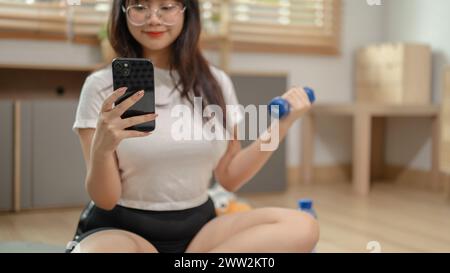 Eine glückliche junge asiatische Frau in Sportbekleidung trainiert mit einer Hantel auf einer Yogamatte im Wohnzimmer, während sie mit ihren Freunden auf ihrem Smartpho plaudert Stockfoto