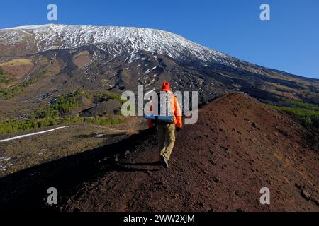 Ein Wanderer mit Rucksack geht auf dem Rücken des erloschenen Vulkankraters im Ätna Park entlang, im Hintergrund schneebedeckten und steilen Hang des Südwestens Stockfoto