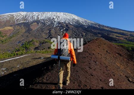 Ein Wanderer mit Rucksack geht auf dem Rücken des erloschenen Vulkankraters im Ätna Park entlang, im Hintergrund schneebedeckten und steilen Hang des Südwestens Stockfoto