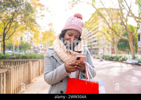 Lächelnde junge Frau, die Einkaufstaschen hält, Smartphone-Apps durchsucht. Stockfoto