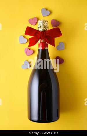 Sektflasche mit Band und hölzernen Herzen auf gelbem Hintergrund gebunden. Festlicher romantischer Hintergrund für Liebhaber Stockfoto