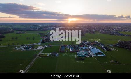 Am Ende des Tages wirft die untergehende Sonne einen strahlenden Hintergrund über eine kleine ländliche Stadt. Diese Luftaufnahme zeigt das Nebeneinander von Kulturfeldern und Wohngebieten, wobei das letzte Licht der Sonne Himmel und Land beleuchtet. Die Abenddämmerung zieht sich über die ländliche Stadt mit dem glühenden Sonnenuntergangshorizont nieder. Hochwertige Fotos Stockfoto