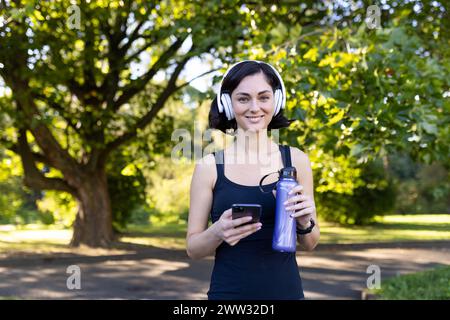 Porträt einer jungen, schönen Frau, die im Park Sport treibt und joggt, in Kopfhörern steht, ein Telefon und eine Flasche Wasser hält. Er schaut mit einem Lächeln in die Kamera. Stockfoto