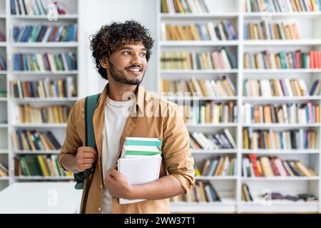 Junger männlicher Student mit lockigen Haaren, die lächeln und Lehrbücher in einer modernen Bibliothek halten, die Selbstvertrauen und Bereitschaft ausstrahlt. Stockfoto