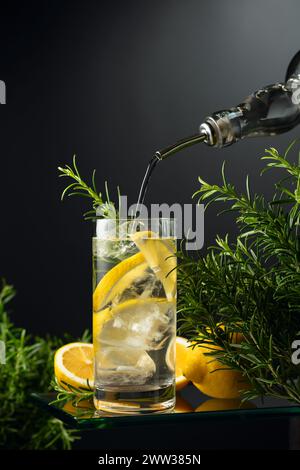 Cocktail-Gin Tonic mit Eis, Zitrone und Rosmarin. Gin wird aus einer Flasche in ein Glas mit Eis- und Zitronenscheiben gegossen. Kopierbereich. Stockfoto