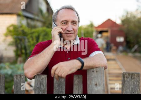 Ein erwachsener Mann, der entspannt neben dem Zaun steht und im Garten telefoniert und lächelt Stockfoto