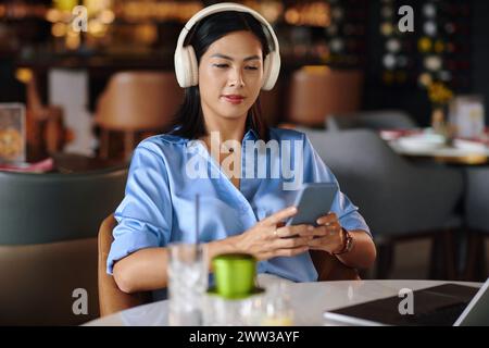 Frau im Kopfhörer sitzt am Restauranttisch und hört Musik auf dem Smartphone Stockfoto