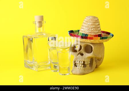 Menschlicher Schädel mit mexikanischem Sombrero-Hut, Tequila und Limette auf gelbem Hintergrund Stockfoto