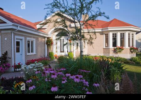 Brauner Ziegelstein mit weißem Zierhaus und landschaftlich gestaltetem Vorhof mit erhöhtem Rand mit rotem Pelargonium, Geranienblumen, Echinacea purpurea, Konefloweren Stockfoto