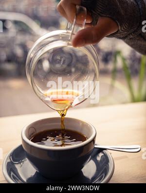 Nahaufnahme einer Hand, die an einem Holztisch in einem gemütlichen Café dampfenden Kaffee in eine Tasse gießt Stockfoto