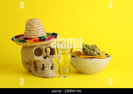 Mexikanischer Sombrero-Hut, menschlicher Schädel, Tequila, Nachos-Chips und Guacamole in der Schüssel auf gelbem Hintergrund Stockfoto