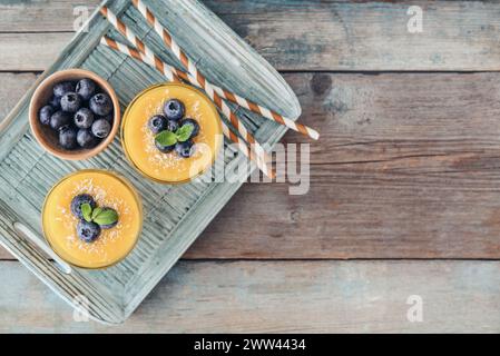 Erfrischender und gesunder Mango-Smoothie in Gläsern mit Kokosflocken und frischen Heidelbeeren auf hölzernem Hintergrund, Blick von oben Stockfoto