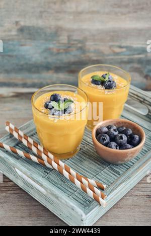 Erfrischender und gesunder Mango-Smoothie in Gläsern mit Kokosflocken und frischen Heidelbeeren auf hölzernem Hintergrund Stockfoto