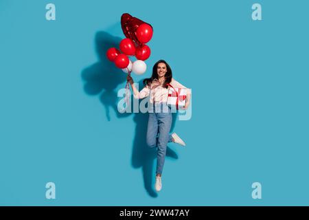 Foto in voller Größe von fröhlicher Frau gekleidetes Seidenhemd, das rote Blasen Geschenkbox in den Händen hält, isoliert auf blauem Farbhintergrund springt Stockfoto