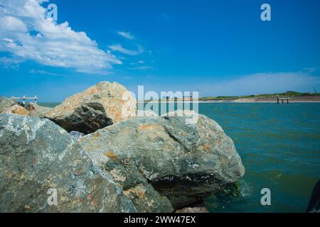 Das Bild zeigt eine friedliche Küste, an der große Felsen als natürliche Barriere zwischen dem üppigen grünen Land und dem ruhigen blauen Wasser dienen. Flauschige weiße Wolken tanzen über den weiten Himmel und spiegeln die Ruhe der Meereslandschaft wider. Coastal Serenity: Rocky Shoreline Trifft Auf Azurblaue Gewässer. Hochwertige Fotos Stockfoto