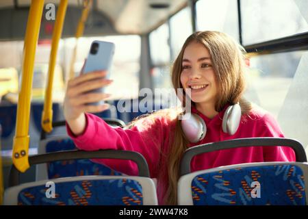 Junges Mädchen in rosa Hoodie und Kopfhörern, das in öffentlichen Verkehrsmitteln, in der modernen Straßenbahn, im Bus sitzt, lächelt und Selfie mit dem Handy macht. Touristen Stockfoto