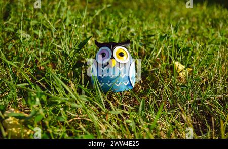 Ein Spielzeug einer überraschten blauen Eule mit mehrfarbigen Augen in Nahaufnahme steht mitten im grünen Gras im Licht der Abendsonne Stockfoto