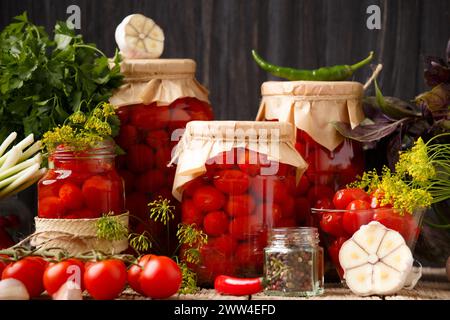 Hausgemachte Tomaten aus der Dose und marinierte Zutaten. Eingelegte Kirschtomaten in geschlossenen und offenen Gläsern, Knoblauch, grüne Zwiebeln, Basilikum und Gewürze Stockfoto