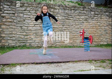 Porträt eines kleinen Mädchens, das auf einem Bein springt und auf einem Spielplatz im Freien Hopscotch spielt. Hopscotch populäres Straßenspiel. Aktives Kind Stockfoto