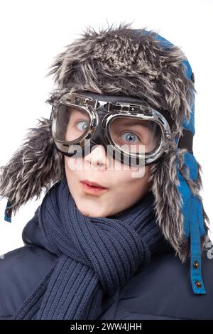 Lächelnder junger Junge, der für Winterspaß gekleidet ist, mit Pilotenbrille und Trapper-Hut aus Kunstfell vor weißem Hintergrund bereit Stockfoto