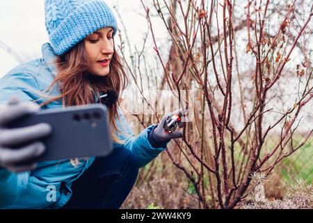 Gärtner-Blogger filmt Vlog über das Beschneiden von Rosenstrauch im Frühlingsgarten. Frau, die mit dem Smartphone Inhalte in sozialen Medien erstellt Stockfoto