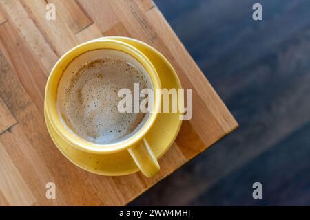 Goldenes Elixier. Gelbe Tasse Macchiato, serviert auf braunem Holztisch, von oben geschossen. Morgendliches Ritual. Stockfoto