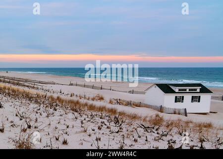 Eine weiße Hütte steht an einem einsamen Strand, mit einem mehrfarbigen Sonnenuntergang Himmel, der eine ruhige Kulisse bietet. Stockfoto