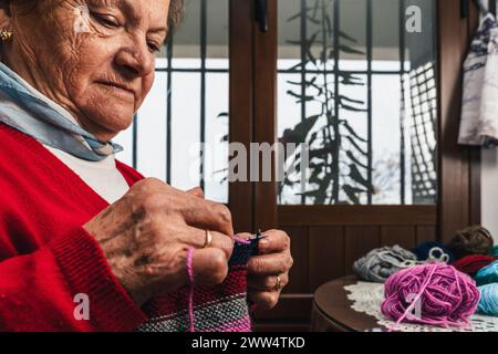 Nahporträt einer hübschen und glücklichen Seniorin, die ein Stück Wollkleidung mit Nadeln und bunter Wolle neben einem Fenster strickt Stockfoto