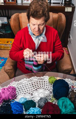 Porträt einer Seniorin und Bewegung ihrer Hände, die mit Nadeln und farbiger Wolle auf einem Sofa zu Hause nähen Stockfoto