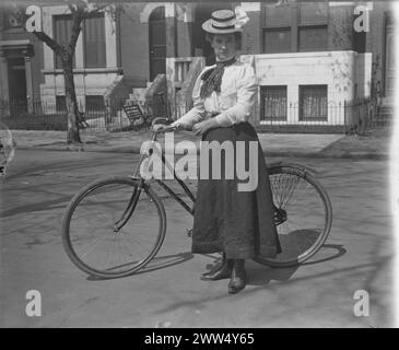 Die Frau trug eine weiße Bluse, einen dunklen Rock und einen Strohhalm mit einer weißen Schleife. Sie steht auf einer gepflasterten Straße, hält ihr Fahrrad, mit einer Reihe von Stadthäusern hinter sich. Schwarz-weiß, ca. 1890er Jahre Foto: Ervin S. Hubbard Stockfoto