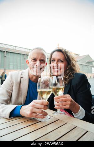 Vertikal. Reifes Paar, das Weißweingläser tröstet, lächelnd und in die Kamera schaut. Verheiratete Menschen mittleren Alters trinken Alkohol auf einer Bar-Terrasse. Freunde im mittleren Erwachsenenalter, die sich mit alkoholischen Getränken anfreunden. Hochwertige Fotos Stockfoto