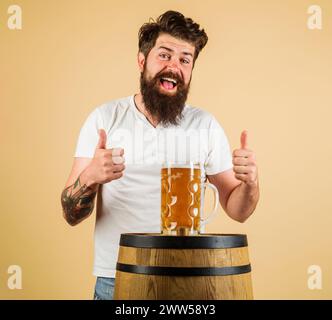 Bärtiger Mann mit einem Becher Bier auf Holzfässern, der Daumen nach oben zeigt. Glücklicher Brauer mit einem Glas Lagerbier. Ausrüstung für die Zubereitung von Bier. Bierkneipe Stockfoto