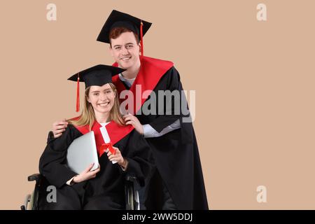 Männlicher Absolvent im Rollstuhl mit Laptop und Klassenkamerad auf beigefarbenem Hintergrund Stockfoto