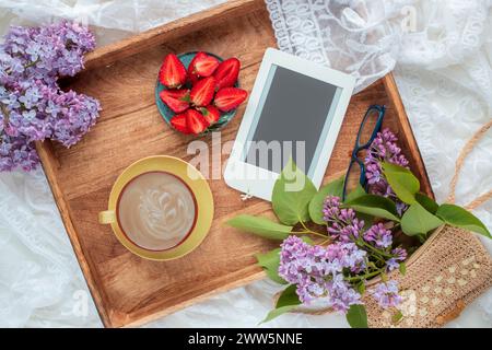 Blick von oben auf Kaffee im Bett mit leckeren Erdbeeren, E-Reader und lilafarbenen Zweigen auf Holztablett. Frühling, Sommer Stillleben Komposition. Stockfoto