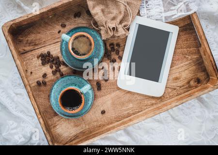 Komposition mit zwei Tassen Espresso, gerösteten Kaffeebohnen und E-Reader mit Kopierraum auf Holztablett im Bett. Stockfoto
