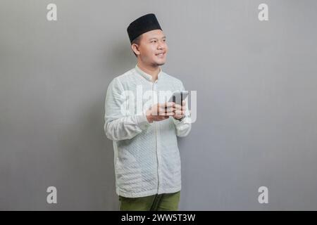Glücklicher asiatischer muslimischer Mann, der Smartphone-Handy für Anrufe, soziale Medien, mobile App online benutzt, ein koko-Shirt und Peki mit Th-Töne trägt Stockfoto