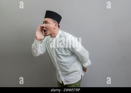 Asiatisch-muslimischer Mann, der die Hände um den Mund schröpft und ein koko-Hemd und Peki mit Farben des Fastenmonats trägt, isoliert auf grauem Hintergrund Stockfoto