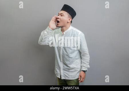 Asiatisch-muslimischer Mann, der die Hände um den Mund schröpft und ein koko-Hemd und Peki mit Farben des Fastenmonats trägt, isoliert auf grauem Hintergrund Stockfoto