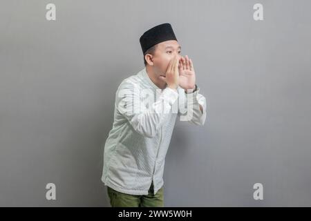 Asiatisch-muslimischer Mann, der die Hände um den Mund schröpft und ein koko-Hemd und Peki mit Farben des Fastenmonats trägt, isoliert auf grauem Hintergrund Stockfoto