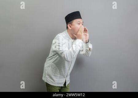 Asiatisch-muslimischer Mann, der die Hände um den Mund schröpft und ein koko-Hemd und Peki mit Farben des Fastenmonats trägt, isoliert auf grauem Hintergrund Stockfoto