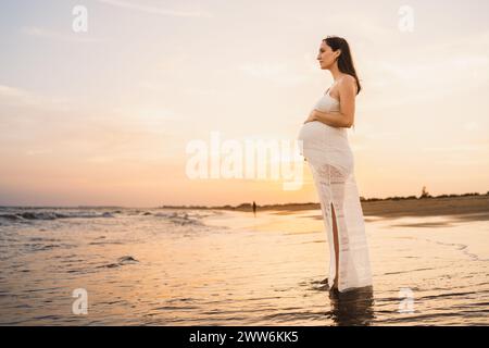 Schöne schwangere Frau in weiß gekleidet, die am Ufer des Strandes steht und auf das Meer blickt Stockfoto