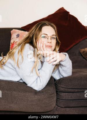 Porträt einer jungen schönen blonden Frau mit Brille, die bequem auf einem Sofa in ihrem gemütlichen Zuhause liegt. Stockfoto