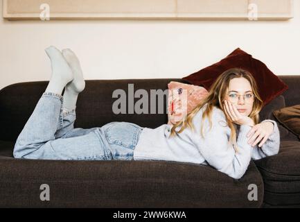 Porträt einer jungen schönen blonden Frau mit Brille, die bequem auf einem Sofa in ihrem gemütlichen Zuhause liegt. Stockfoto