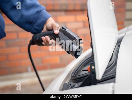 Hand einer Frau, die ein Elektroauto-Ladegerät hält, bereit, das Elektrofahrzeug vor dem Haus anzuschließen. Stockfoto